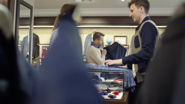 PAN of young man with jacket walking towards checkout counter in clothes shop and chatting with female sales assistant before making purchase 