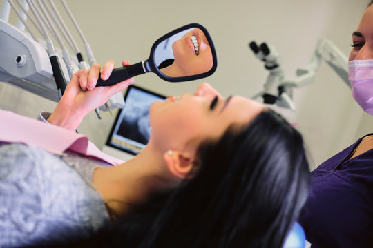 Brunette female sits in a dentist chair and looking into a mirror on her teeth.
