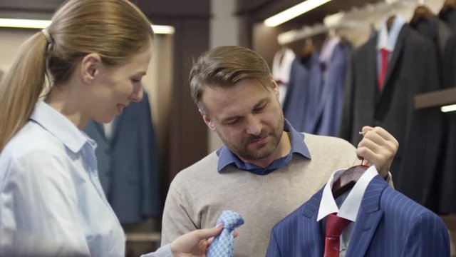 Tilt up of female sales assistant helping male customer choose color of necktie in menswear boutique 
