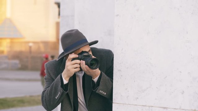 Young male spy agent wearing hat and coat photographing criminal people and hiding behind the wall