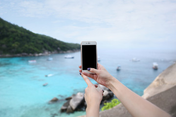 Woman hand hold white mobile phone screen isolated with beautiful fresh sea and blue sky in the background. Similan Islands, Thailand