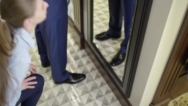 Tilt down with high angle of happy man in tailor made suit checking himself out in mirror while female sales assistant fixing necktie and length of trousers 