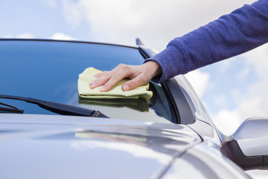 Woman's hand with rag cleaning a silver car's windshield on cloudy sky background in sunny day. Early spring washing or regular wash up. Professional car wash by hands.