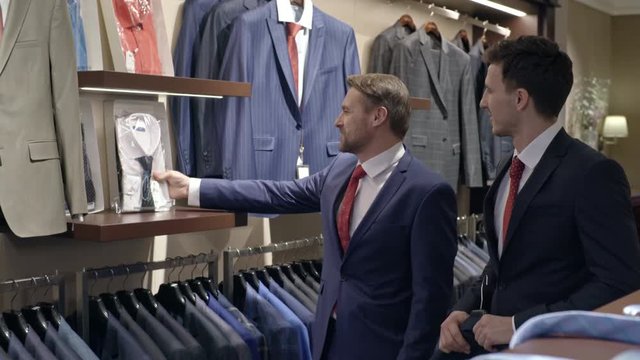 Tracking shot of men in suits walking in menswear boutique and choosing shirt, then asking female customer for advice 