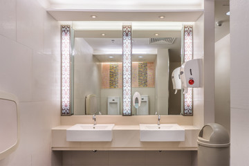 Clean modern restroom with light. It consists of new mirror, ceramic washbasins, granite countertop,liquid soap, plastic bin and toilet paper roll.