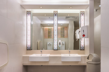 Clean modern restroom with light. It consists of new mirror, ceramic washbasins, granite countertop,liquid soap, plastic bin and toilet paper roll.