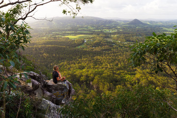 A young man sits, quietly meditating on top of a mountain with expansive views near Noosa Heads,...