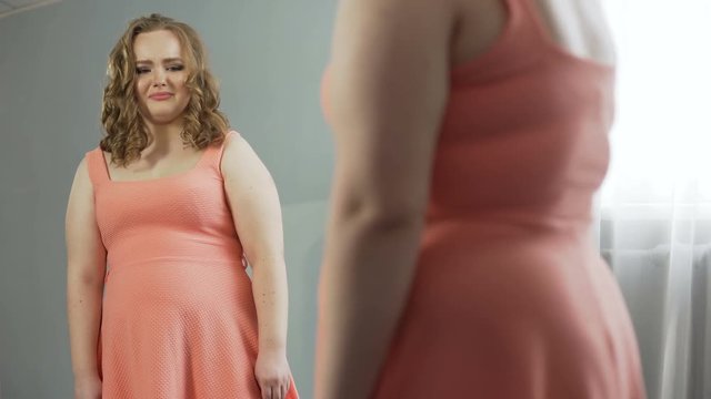 Young woman looking at her mirror reflection with disgust, ashamed of fat body