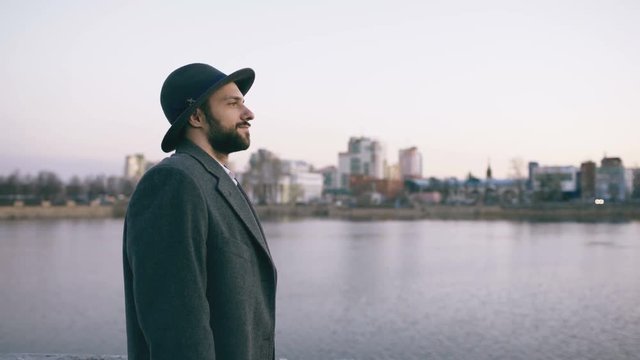 Young bearded tourist man in hat and coat watching cityscape and daydreaming while standing on riverside