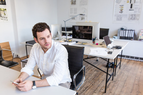 Mid adult man sitting in office at desk holding smartphone looking away
