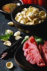 Homemade raw uncooked pasta with whole and sliced organic tomatoes Coeur De Boeuf, salt, seasoning, olive oil and basil over dark wooden background. Italian cuisine