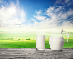 Glass and jug with milk on wooden table on background of pasture