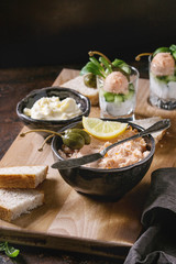 Black bowl of salmon pate with red caviar served with butter, sliced bread, capers, vintage knife, verrines and herbs on wooden serving board, textile linen over brown texture background. Close up