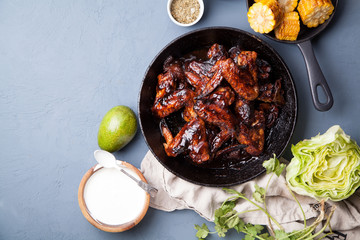 Chicken wings in thick barbeque sauce with grilled corn, sour cream sauce, honey and salad in black cast iron pan on gray rustic background. Mexican cuisune. Copy space.