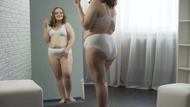 Body positive girl happy with her weight, devouring donuts in front of mirror