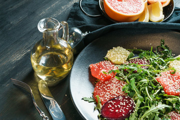Composition from a bottle with olive oil and a black dish with fresh salad from red beet, grapefruit and orange on a cushion of arugula on a textured wooden table top in black.