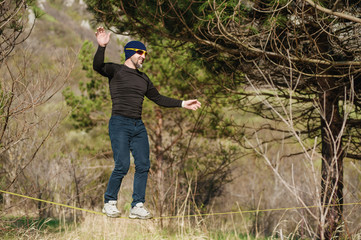 A man, aged with a beard and wearing sunglasses, balances on a slackline in the open air between two trees at sunset