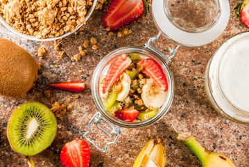 Healthy breakfast. Diet. Overnight oatmeal in a can, muesli. Yogurt with homemade granola and organic fruits - kiwi, banana, strawberry. On the stone table. With ingredients and spoons. 