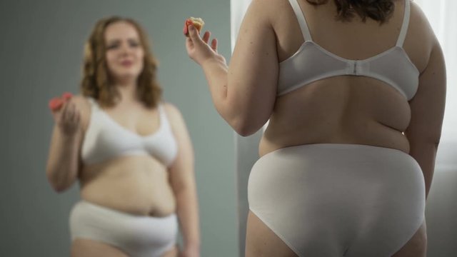 Overweight young woman stress eating in front of mirror, hating her fat body