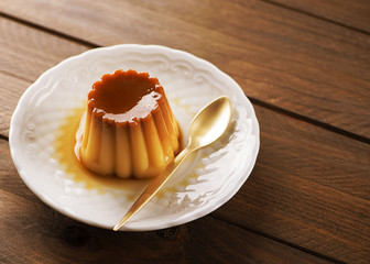 From above white plate with flan and spoon on the wooden background.