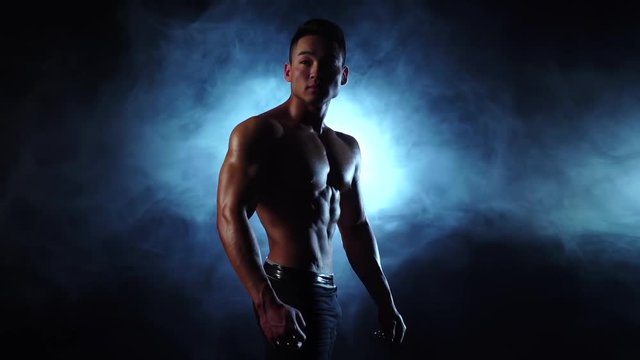 Asian bodybuilder man demonstrates his body, strength and endurance. Black smoke background. Slow motion
