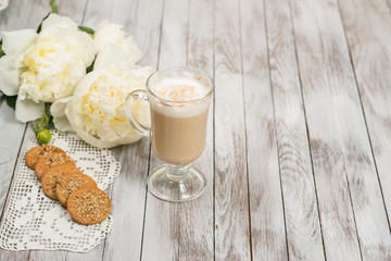Glass of cappuccino next to cookies and pionies flowers on white wooden background. Place for text.