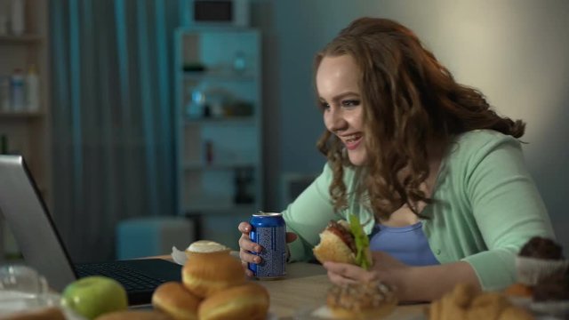 Teenager girl enjoying comedy on laptop drinking soda and eating burger meantime