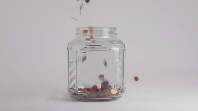 Coins Fall into Clear Jar in Slow Motion. a clear jar fills up with coins dropped from above, spilling and filling the jar in slow motion.

