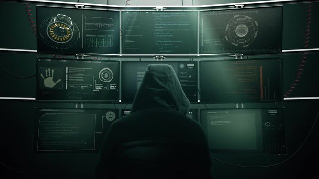 Hooded Man Watches Hacker Monitors. A man in a hooded sweatshirt watches a variety of video monitor screens on a wall display various data and surveillance images 
