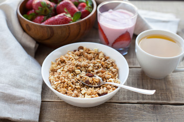 A bowl of homemade granola with yogurt and fresh strawberries on a wooden background. Healthy breakfast with green tea