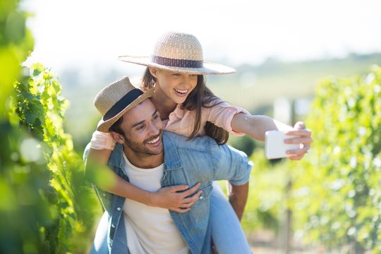 Happy couple taking selfie while piggybacking at vineyard - Powered by Adobe