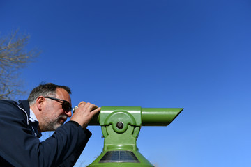 Ein Älterer Mann schaut bei klarem blauem Himmel durch ein festmontiertes grünes Fernglas 