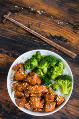 A spicy, homemade teriyaki chicken with sesame seeds in a white bowl, garnished with rice and broccoli. Wooden rustic table, top view.