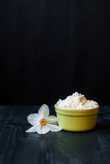 Just cottage cheese in ceramic ware on a dark background. Sour grainy cheese on a wooden table. Curd and daffodil flower.