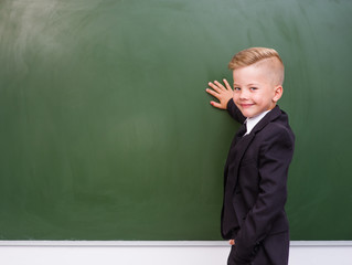 Happy boy in a suit standing near empty green chalkboard