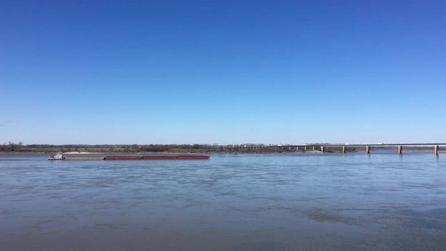 Tugboat pushes a barge up the Mississippi River toward Interstate 40 next to Memphis, Tennessee with Arkansas in the background.