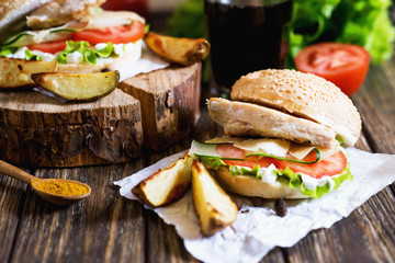Fast food, barbecue, burger with a white bun with sesame, grilled chicken fillet, fresh cucumber, tomato, lettuce, mayonnaise, with a glass of cola and baked potato on a dark wooden background 