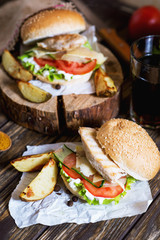 Fast food, barbecue, burger with a white bun with sesame, grilled chicken fillet, fresh cucumber, tomato, lettuce, mayonnaise, with a glass of cola and baked potato on a dark wooden background 