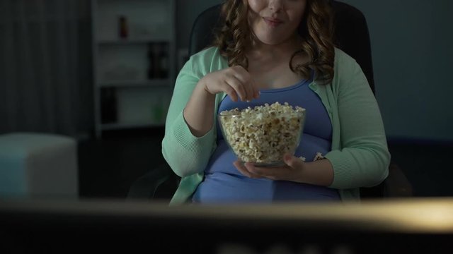 Young chubby woman laughing, commenting on TV show and eating popcorn messily 