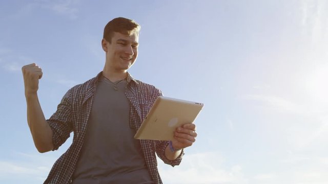 Joyful toung man is tallking with someone over his tablet pc while standing on a beach