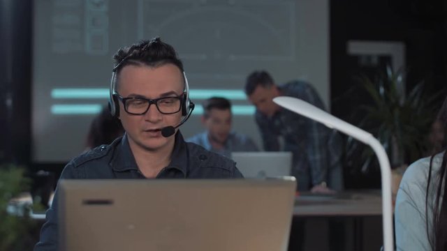 Men with headset working in call center in modern worldwide office he talking and looking on screen of desktop computer solving the client's problem online.