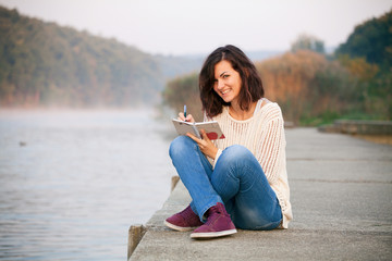 Writing in a notebook. Happy woman is writing her diary in the morning. Sitting by lake.