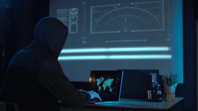 Man in the hood from his back sitting and working at laptop as hacker, with world map on wall in background. Then he take off usb flash disk and run away. Perhaps he has stolen information or has