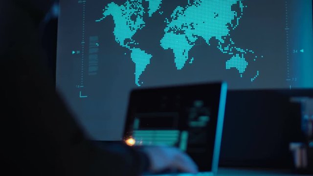 Hands of unrecognizable man hacker typing a laptop in futuristic modern office with animated world map on background