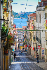 Fototapeta na wymiar Steep narrow street of Lisbon with cable car rails in the middle and ocean in the background