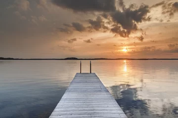 Papier Peint photo Lavable Jetée Beautiful glowing orange sunset over a rustic timber plank jetty reflected in the mirror calm waters of the sea below, a background of natural beauty and serenity. Northern sea, Sweden, Scandinavia.