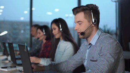 Young handsome men in checked shirt and headset working in call center in modern worldwide office he talking and looking on screen of desktop computer