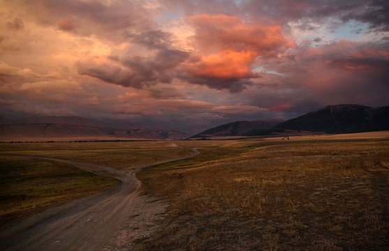Road path on a desert wild mountain plateau at the background of the hills under a dramatic sunset colorful sky with illuminated red pink purple clouds Kurai Altai Mountains Siberia Russia