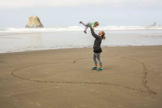 Mother lifting daughter on heart shape outline on sand