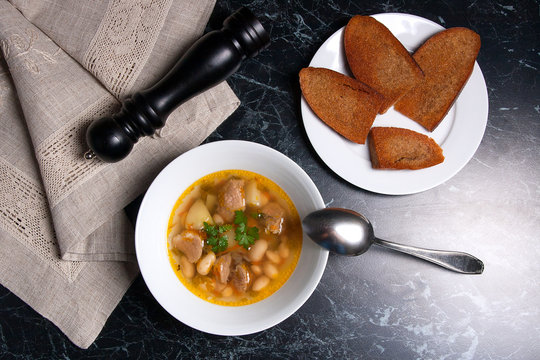 Bean soup in white plate with metal spoon, several toast on white plate on a black stone background.